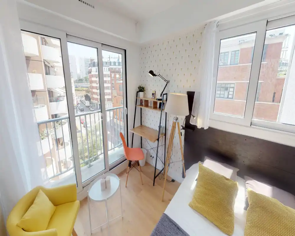 Camera da letto luminosa e compatta con letto matrimoniale, cuscini decorativi e un piccolo angolo lavoro vicino alla finestra. Ampie porte scorrevoli e una seconda finestra garantiscono molta luce naturale e vista sulla strada.