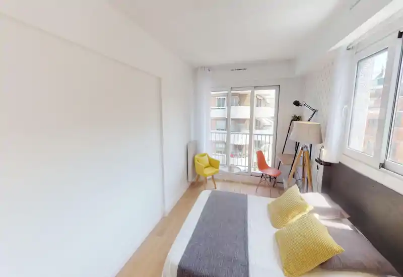 Camera da letto luminosa e moderna con un letto ampio, cuscini gialli come accento, una piccola area salotto vicino a grandi finestre e un angolo scrivania compatto — ideale per un affitto cittadino soleggiato.
