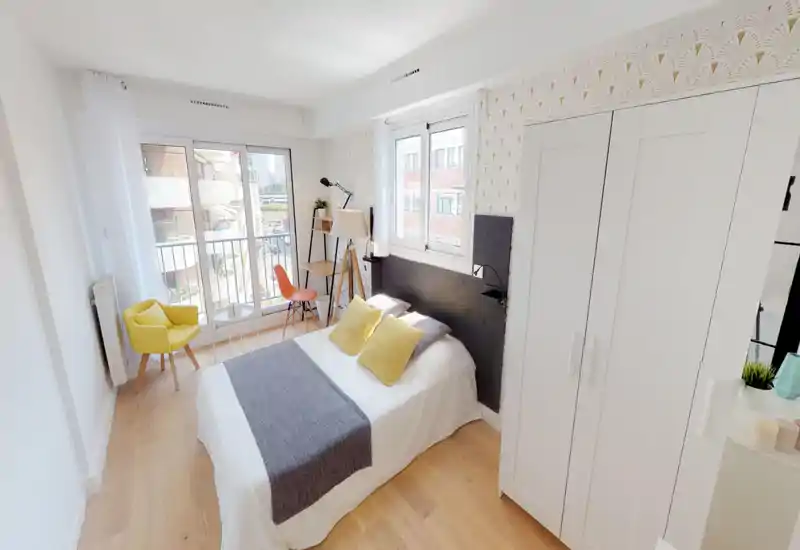 Camera da letto luminosa e ben arredata con letto matrimoniale in lenzuola bianche, runner grigio e cuscini gialli. Ampie finestre e porta-balcone illuminano la stanza; un armadio bianco e un'area salotto completano lo spazio.