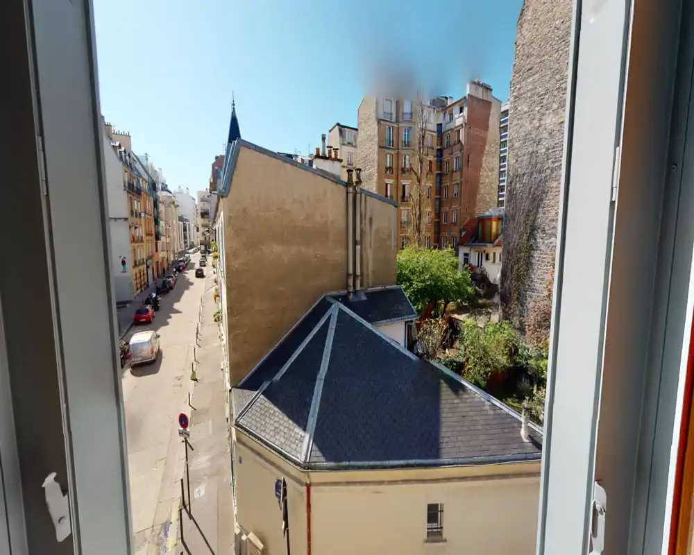 Bright urban view from a window showing neighboring rooftops, a quiet street and courtyard greenery — ideal for highlighting building location and light in a listing.