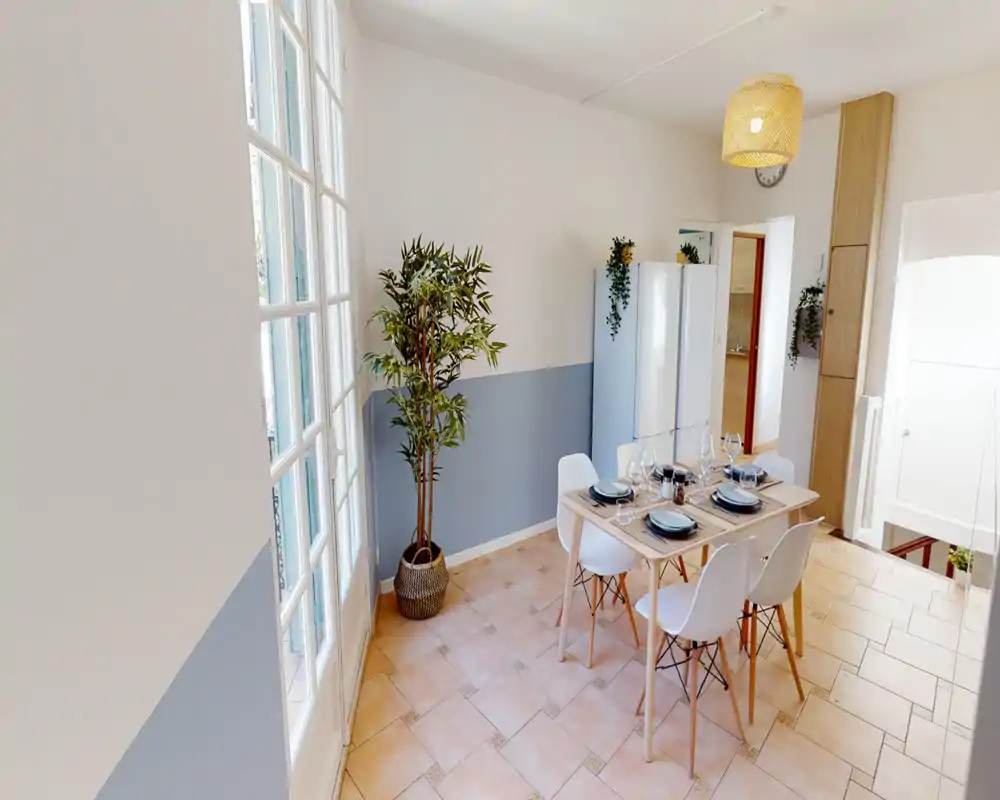 Bright, modern dining area with a set table for four, light wood table and white chairs beside tall windows; stylish pendant lamp and potted plant add warmth.