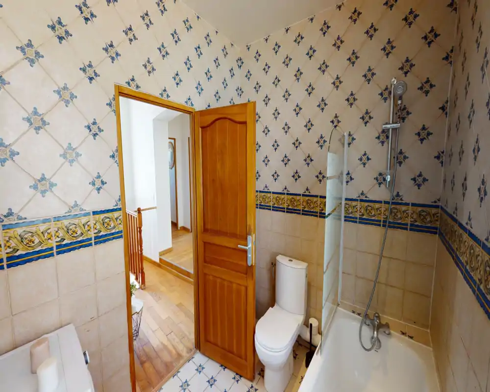 Tiled bathroom with patterned ceramic walls, a bathtub with showerhead and glass screen, and a toilet next to a wooden door leading to a hallway.
