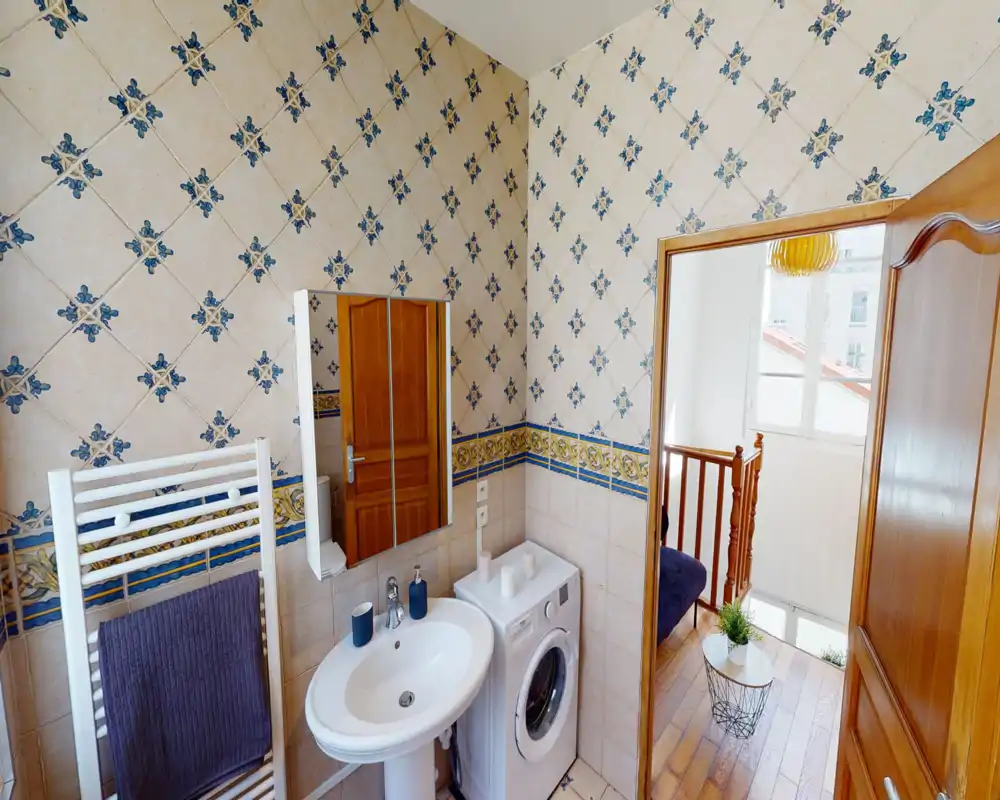 Bright tiled bathroom with a pedestal sink, mirror cabinet, towel warmer and a washing machine; patterned blue-and-cream tiles and good natural light from a nearby stairwell window.