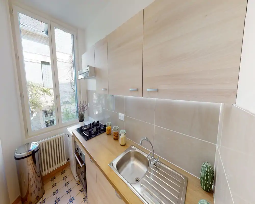 Bright, compact kitchen with wooden cabinets, stainless steel sink and gas hob next to a large window — clean and well-styled for a city apartment.