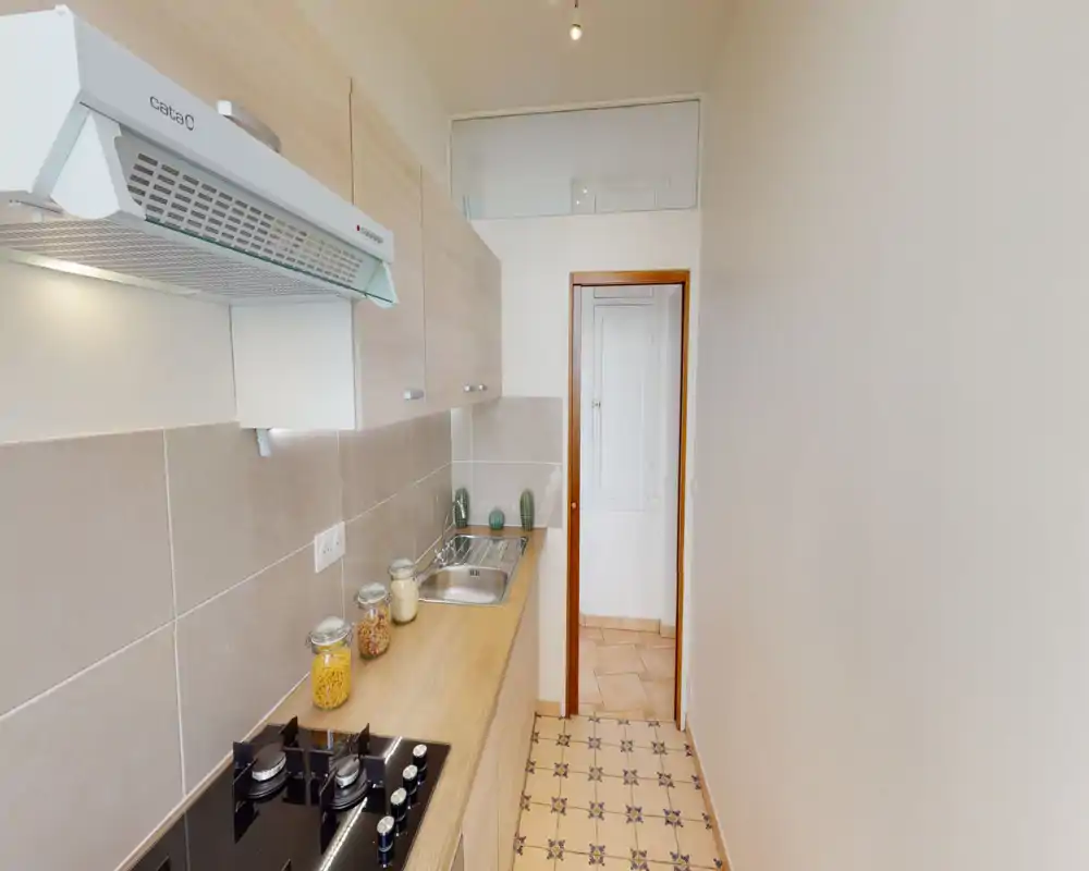 Compact galley kitchen with a modern gas hob, stainless sink, light wood countertops and upper cabinets. Decorative jars and small plants give a neat, inviting feel.