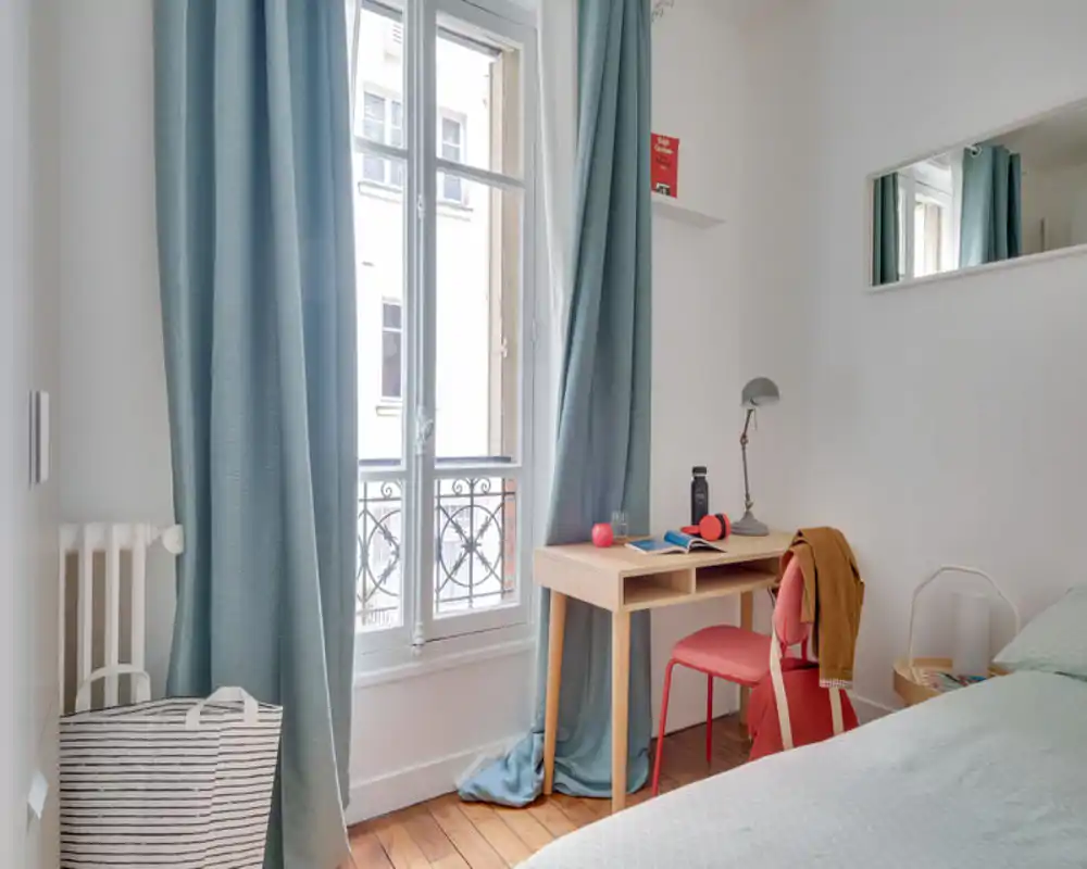 Bright, neatly staged bedroom corner with a visible bed, large window with teal curtains, and a small wooden desk by the window — ideal for a cozy rental listing.