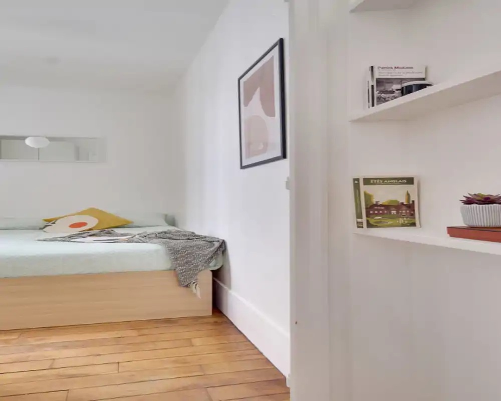 Bright, minimalist bedroom corner with a low wooden bed, soft pastel bedding, a decorative cushion and a cozy throw. The composition shows calm, Scandinavian-inspired styling and natural light.