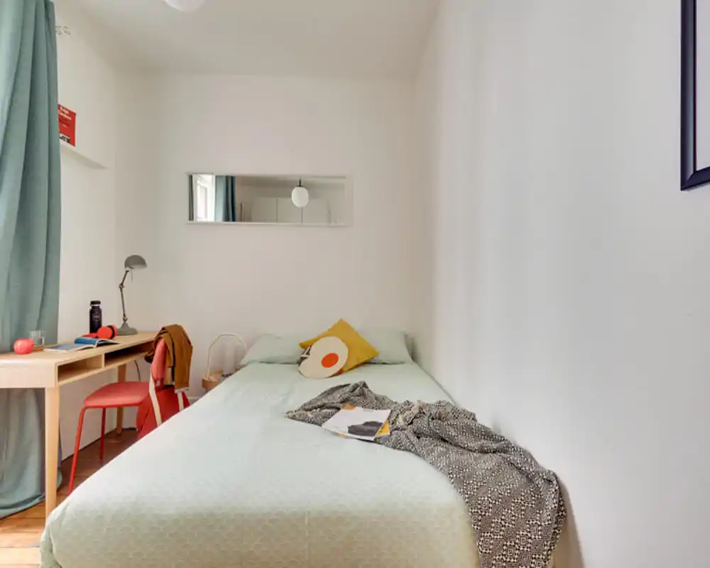 Compact, well-styled single bedroom with a large bed occupying the foreground, soft pastel bedding, decorative pillows and a cozy throw. A small desk by the window provides a practical workspace with natural light.