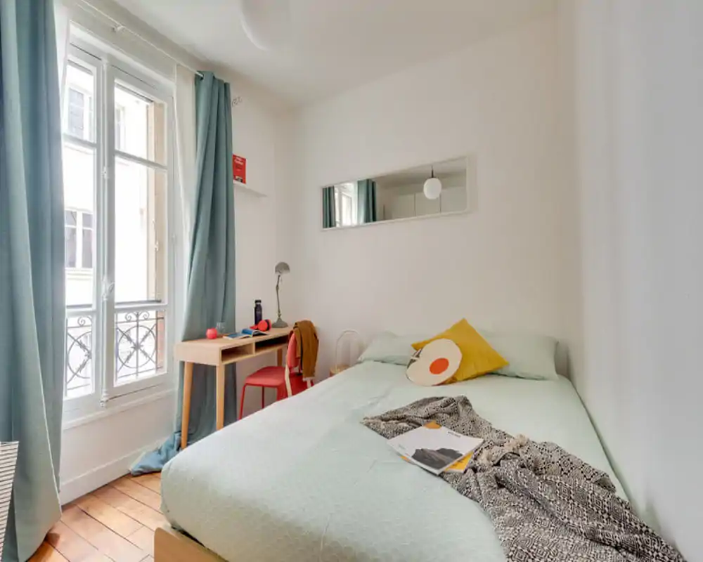 Bright, well-styled bedroom with a double bed, soft mint bedding, decorative cushions and a small desk by a large window letting in abundant natural light.