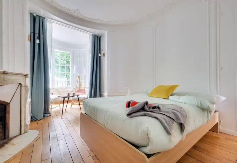 Chambre lumineuse et élégante avec un grand lit sur structure en bois au premier plan et un coin salon baigné de lumière près de la fenêtre en baie. Moulures classiques et parquet chaud pour une ambiance raffinée et accueillante.