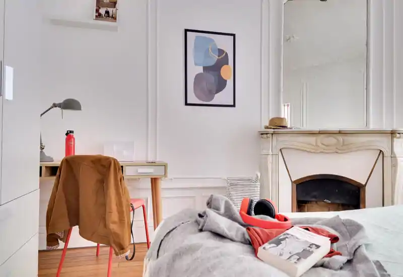 Chambre lumineuse et stylée avec lit au premier plan, cheminée décorative et un petit bureau sur le côté — un espace chaleureux et bien éclairé idéal pour une annonce locative.