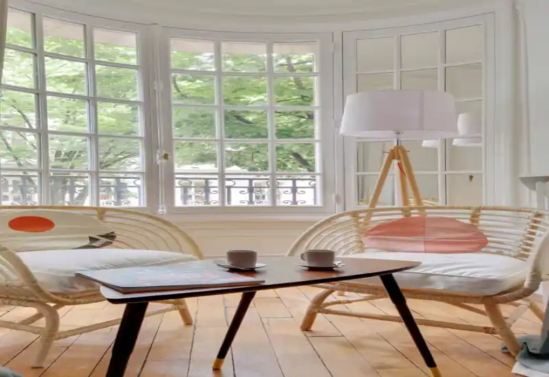 Espace de vie lumineux et chaleureux avec deux fauteuils en rotin, une petite table basse en bois avec des tasses et une grande fenêtre cintrée laissant entrer beaucoup de lumière naturelle.
