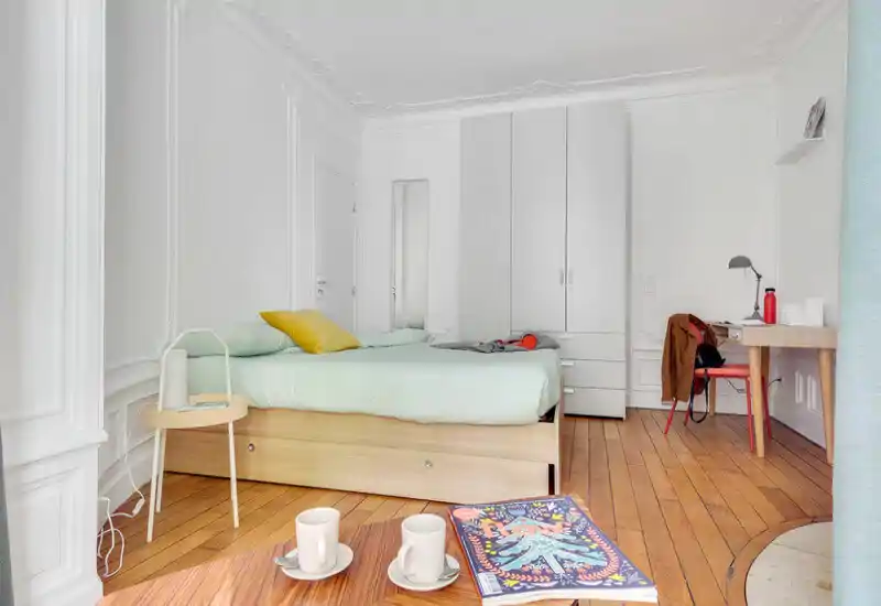 Chambre lumineuse et bien décorée avec un grand lit, linge clair et parquet en bois ; placard et bureau ajoutent du confort pour un séjour en ville.