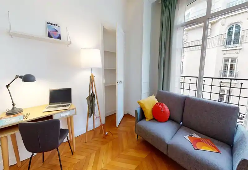 Espace de vie moderne et lumineux avec un petit canapé gris, des coussins colorés et un bureau près de la fenêtre. Les grandes fenêtres et le parquet chaleureux rendent la pièce accueillante et aérée.