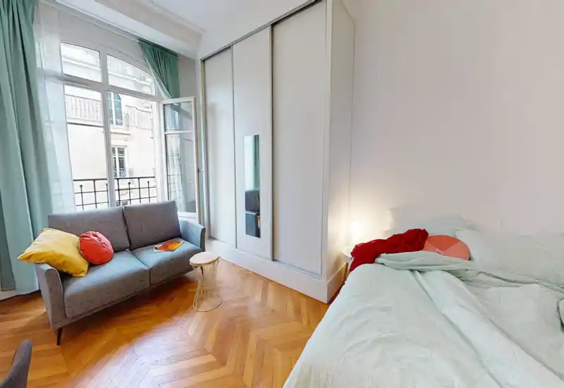 Chambre lumineuse et chaleureuse avec un lit double confortable, linge de lit vert menthe et un petit coin salon près d'une grande fenêtre laissant entrer beaucoup de lumière naturelle.