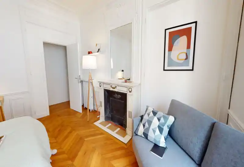 Camera da letto luminosa ed elegante con parquet a spina di pesce, camino decorativo, grande specchio e un piccolo divano accanto al letto — ambiente caldo e curato, ideale per annunci.