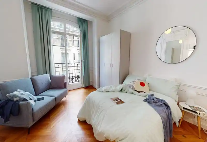 Camera da letto luminosa e di stile con letto matrimoniale, cuscini decorativi, uno specchio rotondo a parete e un piccolo divano accanto a una finestra ad arco che illumina con luce naturale.