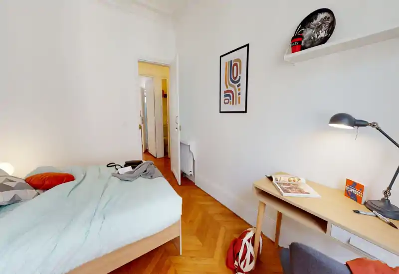 Chambre lumineuse et chaleureuse avec un cadre de lit en bois, une literie douce et un petit bureau avec lampe. Le parquet et les cadres muraux simples donnent une atmosphère calme et bien éclairée, idéale pour se reposer ou travailler.