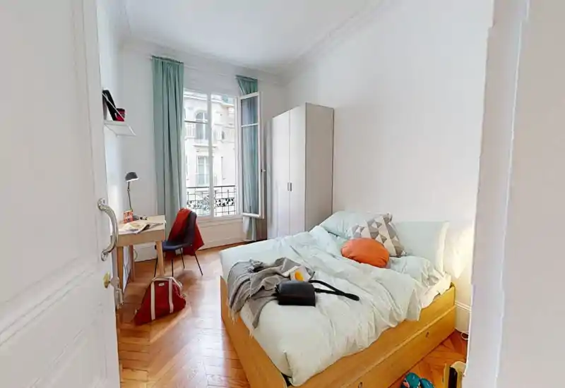 Chambre lumineuse et chaleureuse avec un grand lit, literie pastel et coussins décoratifs. Une armoire, un bureau et une fenêtre avec rideaux clairs complètent cet espace aéré et bien éclairé.