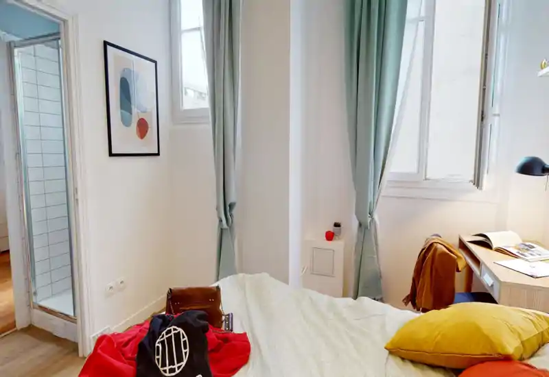 Camera da letto luminosa e curata con un ampio letto in primo piano, tende verde chiaro e una scrivania vicino alla finestra. Tessuti caldi e luce naturale la rendono ideale per annunci.