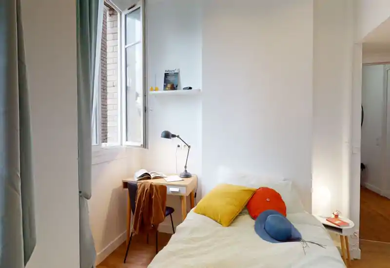 Piccola camera da letto luminosa con letto singolo decorato con cuscini colorati, una lampada da comodino e una scrivania compatta vicino alla finestra che lascia entrare luce naturale.