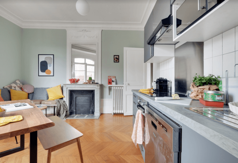 Espace de vie lumineux et élégant, intégré à une cuisine moderne compacte. Murs aux tons pastels, canapé avec coussins colorés et table en bois offrent une atmosphère chaleureuse et accueillante, idéale pour se détendre ou recevoir.