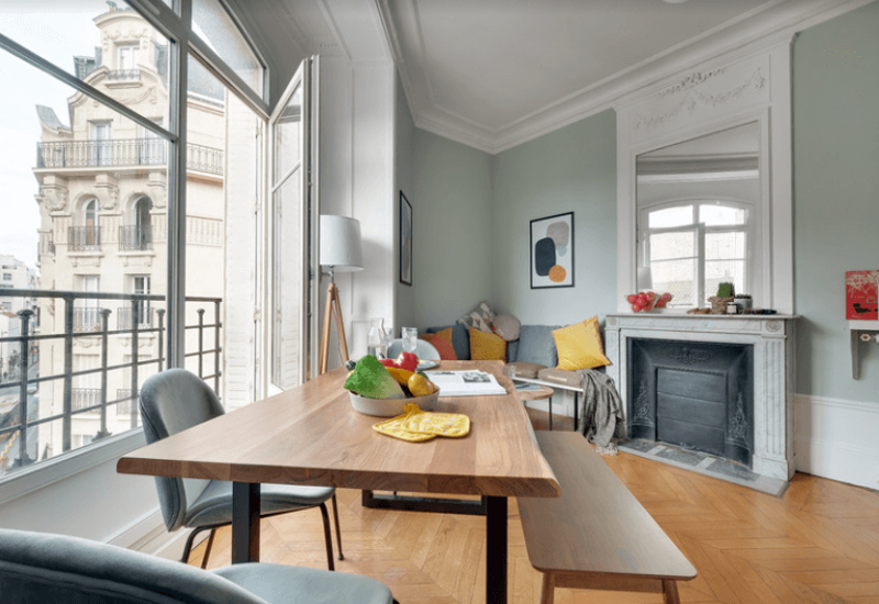 Salon lumineux et élégant avec table en bois, assises confortables, coussins décoratifs et grande fenêtre donnant sur des façades classiques — parfait pour dîner et se détendre.