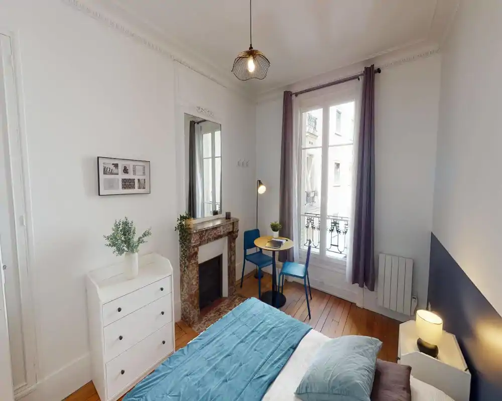 Chambre lumineuse et soignée avec couvre-lit bleu, lampe de chevet et petite table près de la fenêtre. La pièce dispose d'une cheminée décorative, d'un grand miroir et de parquets, créant une atmosphère chaleureuse et bien éclairée.