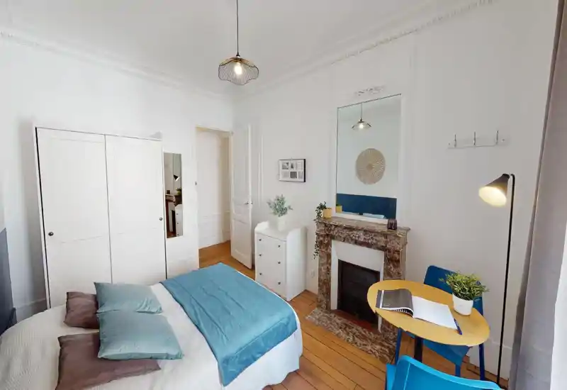 Chambre lumineuse et bien mise en scène avec un lit double aux draps bleus, parquet, une cheminée décorative et une petite table près de la fenêtre — aérée et accueillante.