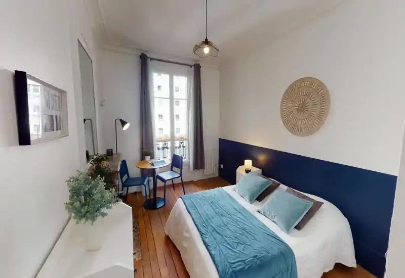 Chambre lumineuse et soignée avec un lit double habillé de textiles bleu-vert, une table de chevet avec lampe, une petite table près de la fenêtre et un parquet chaleureux. Grande fenêtre apportant une belle lumière naturelle et décoration harmonieuse.