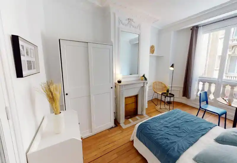 Sunny, stylish bedroom with a comfortable double bed dressed in blue linens, classic fireplace with mirror, wardrobe and a balcony letting in abundant natural light.