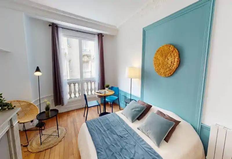 Bright, stylish bedroom with a queen-size bed against a turquoise feature wall, soft linens and decorative cushions, plus a small dining table by a large window letting in generous natural light.