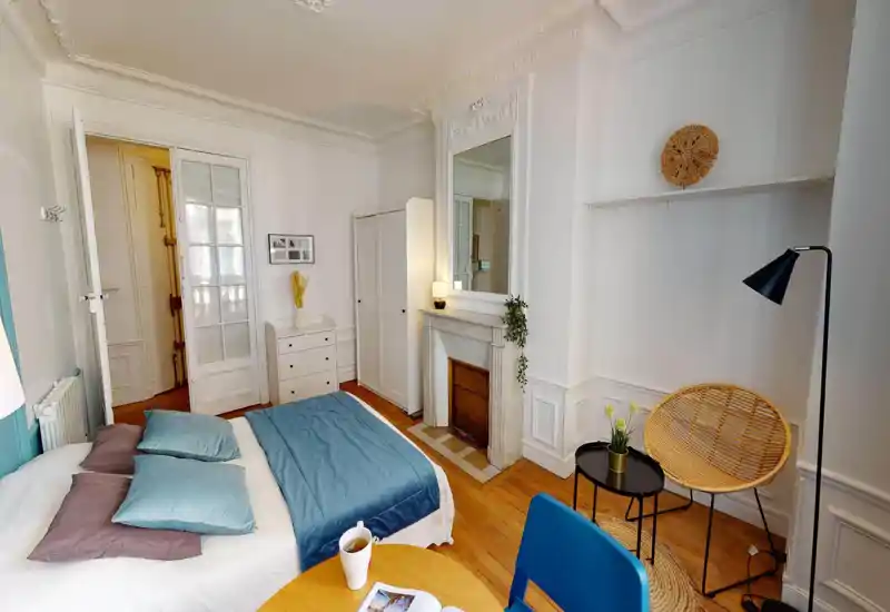 Bright, elegant bedroom with a double bed dressed in blue linens, decorative pillows, a dressing mirror above a decorative fireplace, wardrobe and cozy seating area — ideal for comfortable stays.