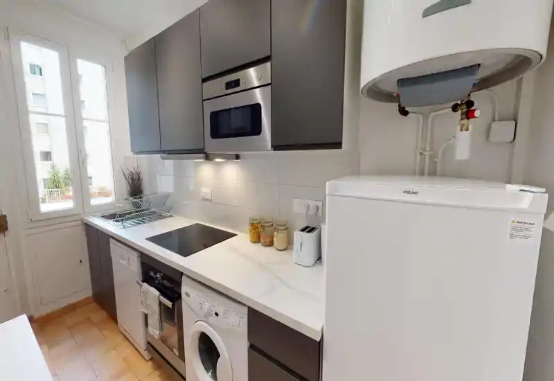 Cuisine moderne et compacte avec plan de travail effet marbre, four encastré, plaque électrique et grand réfrigérateur. La lumière naturelle par la fenêtre et le rangement soigné rendent cet espace lumineux et fonctionnel pour un petit appartement.