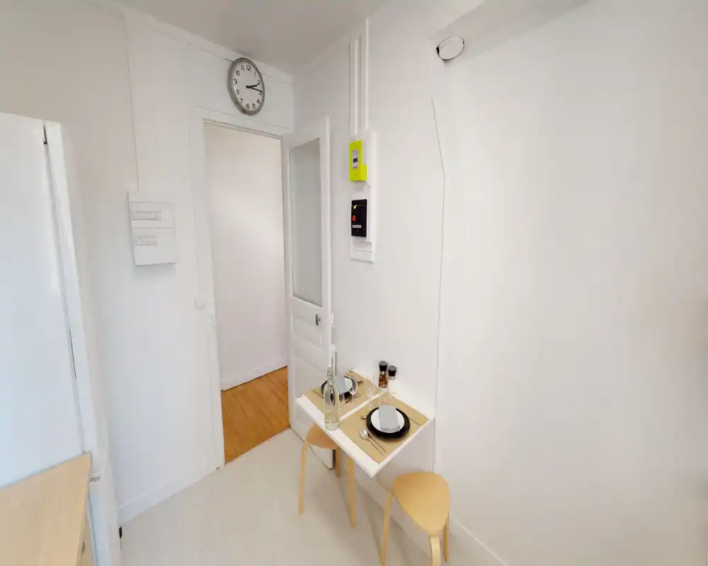 Compact, bright kitchen area with a small fold-down dining table set for two, light wooden stools and minimalist white finishes.
