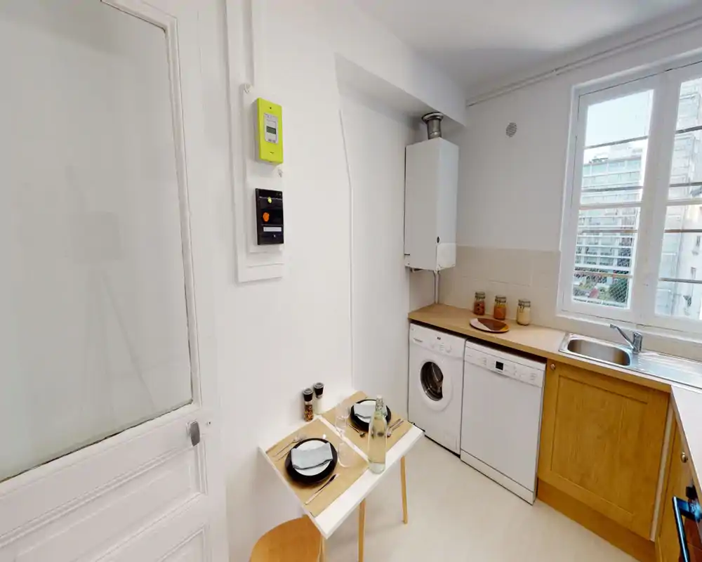 Compact, bright kitchen with wooden cabinets, sink by a large window, dishwasher and washing machine under the counter, and a small two-place dining table.