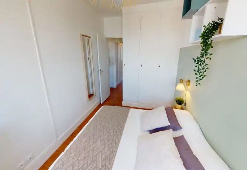 Chambre lumineuse et bien aménagée avec un grand lit au premier plan, coussins doux et une lampe murale. Tons neutres et plante suspendue pour une ambiance calme et accueillante, idéale pour une annonce locative.