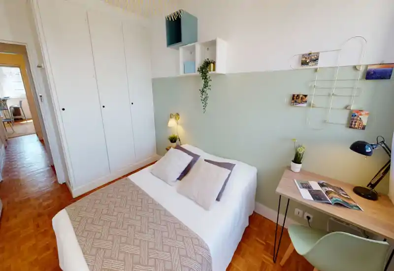 Chambre lumineuse et accueillante avec lit double bien fait, lampe de chevet et petit bureau — mur ton menthe et placards intégrés pour un espace à la fois pratique et élégant.