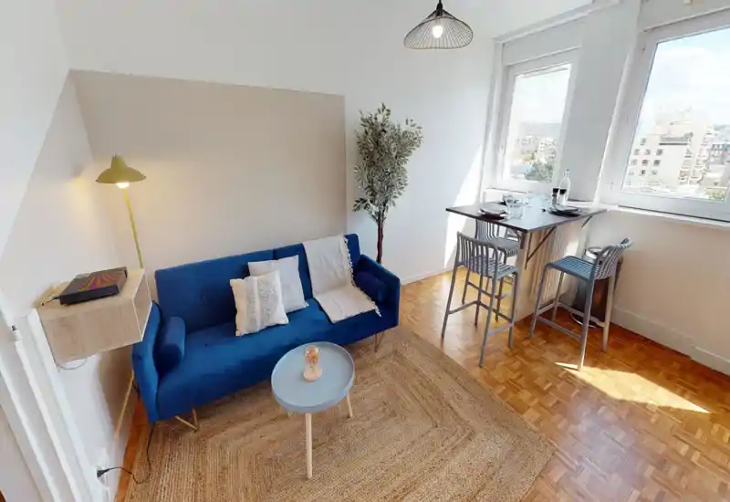 Espace de vie lumineux et soigné avec un canapé bleu, tapis tressé et un petit bar repas près des fenêtres — parfait pour un appartement citadin.