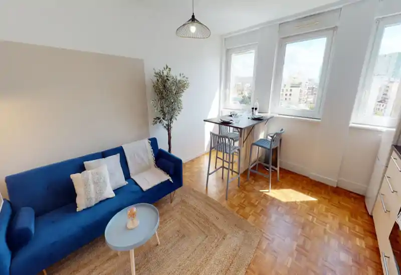 Sala luminosa y con estilo con sofá azul, alfombra tejida y una pequeña barra comedor junto a ventanas soleadas; ideal para un piso urbano compacto.