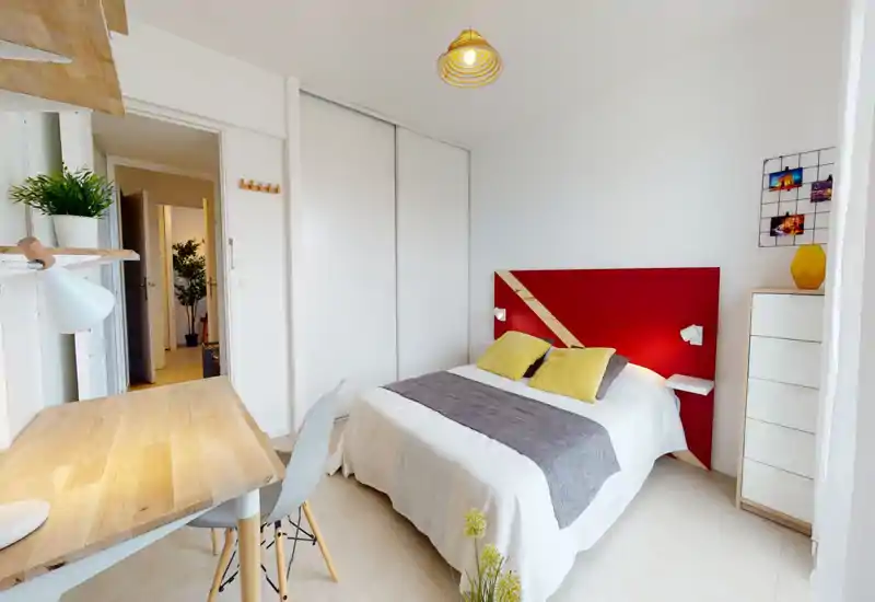 Chambre lumineuse et moderne avec un lit double, une tête de lit rouge et des coussins jaunes. La pièce comprend un petit bureau, une armoire coulissante et une décoration claire et aérée, idéale pour la location courte durée.