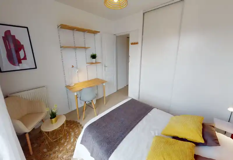 Chambre lumineuse et bien décorée avec un grand lit au premier plan, coussins jaunes en accent et un tapis chaleureux ; un petit bureau et des étagères la rendent pratique pour le télétravail.
