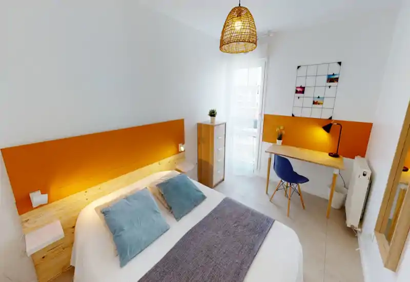 Chambre moderne et lumineuse avec lit double, tête de lit orange et petit bureau près de la fenêtre ; décorée de coussins bleus et baignée de lumière naturelle.