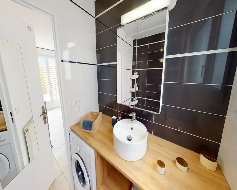 Coin salle de bain moderne avec vasque ronde posée sur un plan de travail effet bois et un carrelage foncé en arrière-plan. Armoire miroir, robinet et petits accessoires visibles, une machine à laver est installée sous le plan.