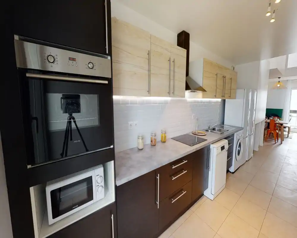 Cuisine moderne et entièrement équipée avec four encastré, micro-ondes, plaque à induction, évier et éclairage intégré. Les meubles hauts en bois clair et les éléments bas foncés offrent un contraste élégant ; plan de travail dégagé.