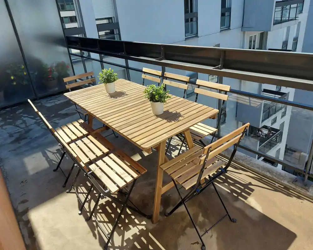 Balcon ensoleillé avec table à manger en bois et six chaises pliantes assorties, deux petits pots sur la table et garde-corps en verre/métal donnant sur les immeubles voisins — idéal pour les repas en extérieur.