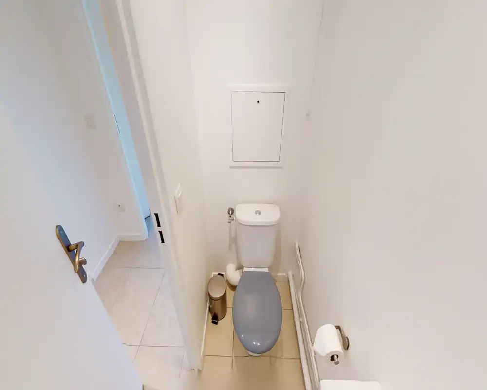 Petite salle de bains montrant de près les toilettes avec abattant gris, une petite poubelle à pédale et un sol carrelé. Murs blancs lumineux et équipements simples pour un rendu propre et minimaliste.