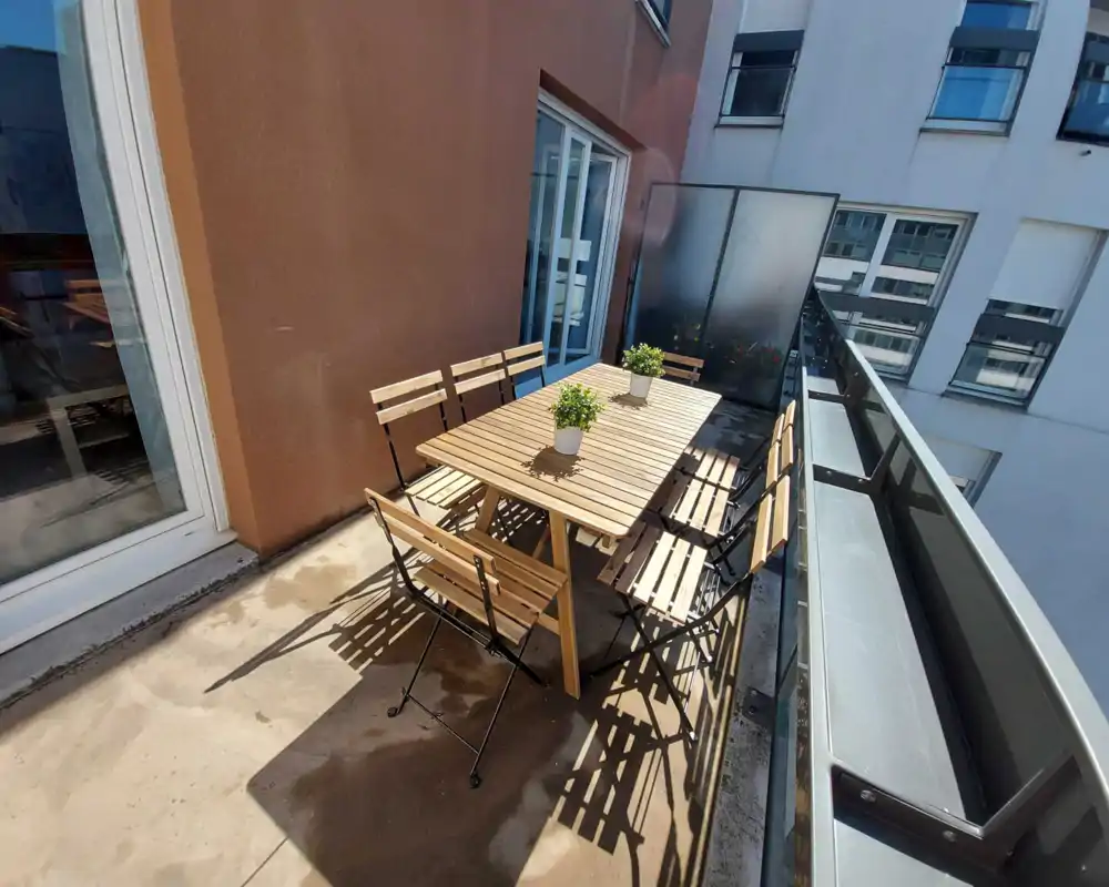 Balcon ensoleillé et étroit avec une table à manger en bois et des chaises pliantes, deux petites plantes en pot et une rambarde en verre — parfait pour repas en extérieur ou café.