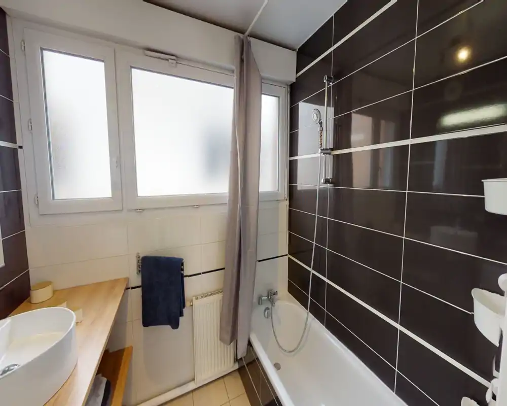 Salle de bains moderne et lumineuse avec baignoire et douche, carrelage mural sombre à l'horizontale et fenêtre dépoli pour la confidentialité.