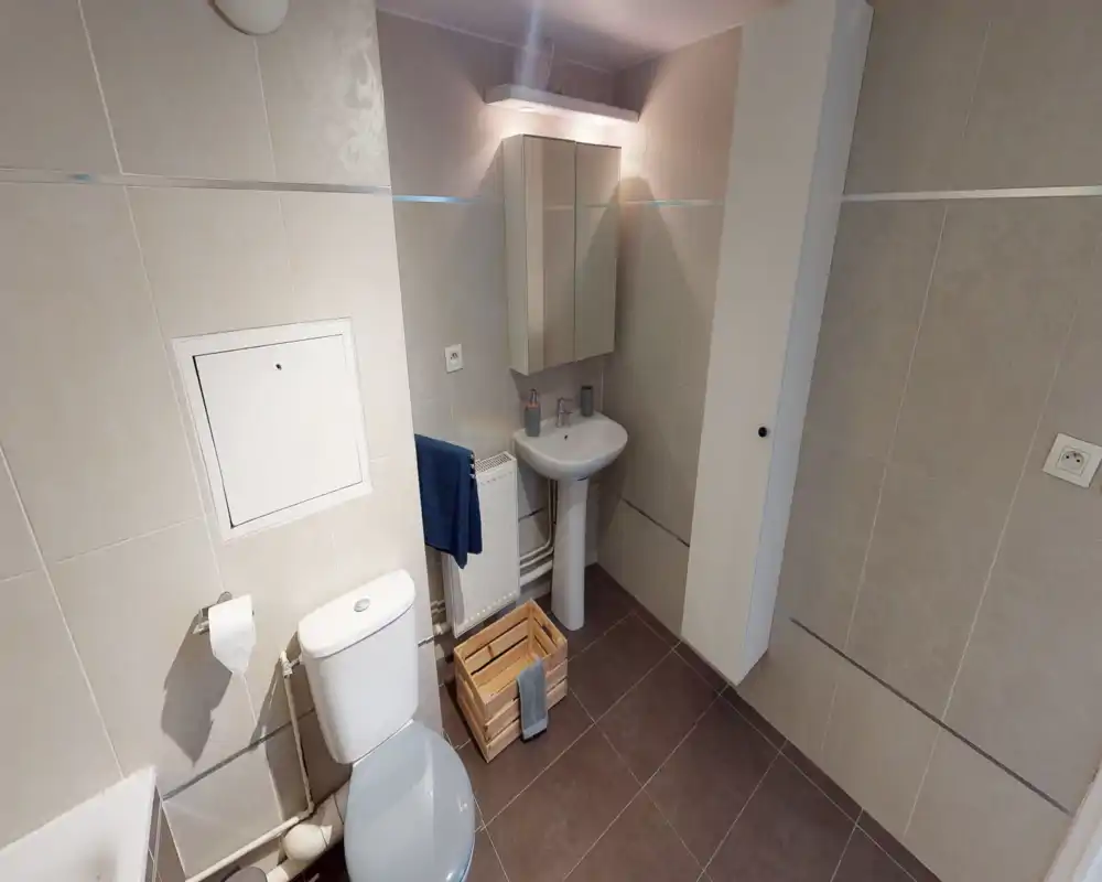 Salle de bains compacte et moderne avec lavabo sur colonne blanc, WC, armoire-miroir et murs carrelés ; propre et bien éclairée, adaptée à une annonce locative.